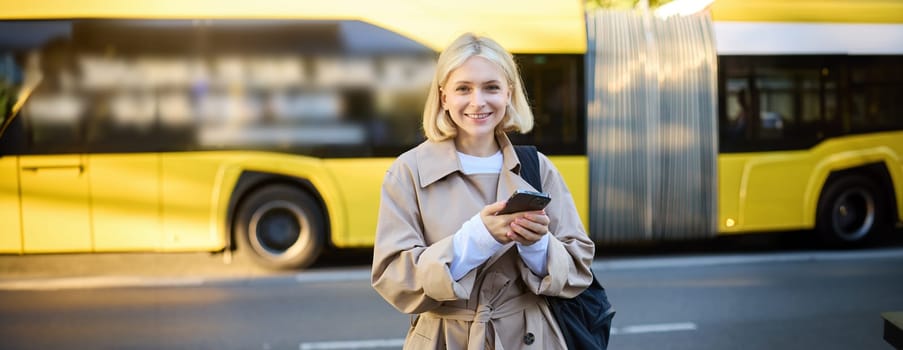 Outdoor shot of young woman with backpack, standing on street with cars passing behind her, holding mobile phone and smiling, urban lifestyle concept.