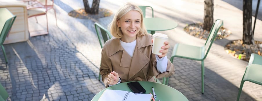 Beautiful blond woman, smiling, showing white takeaway coffee cup, drinking beverage in cafe shop, sitting outdoors.