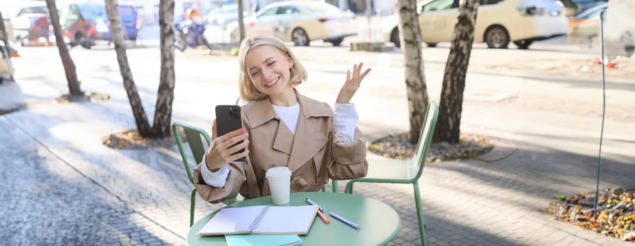 Young blond female model, girl student in cafe, discuss something over the phone, video chatting, looking at smartphone camera and smiling, talking to friend over mobile app.