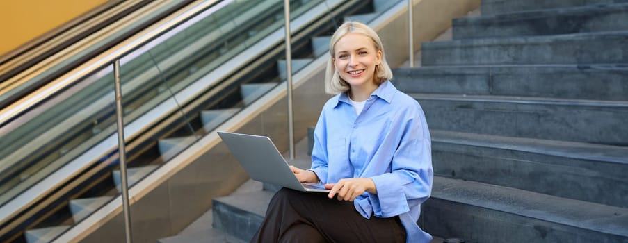 Smiling girl student using laptop computer modern technology device outdoor in university campus online learning, elearning outside sitting on urban stairs. Web education course webinars concept.