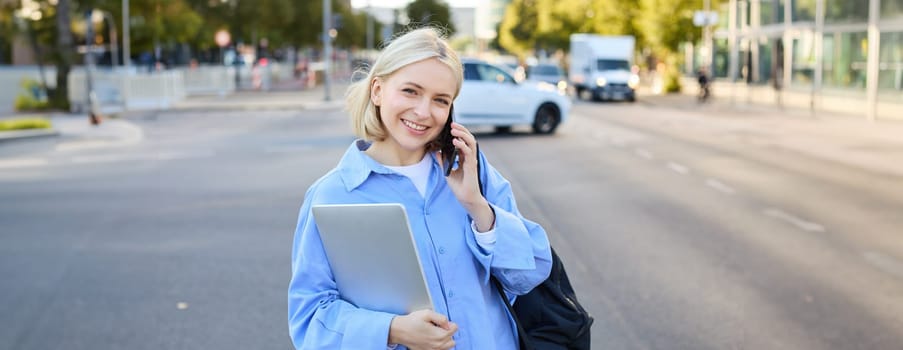 Portrait of young modern female model in blue shirt, talking on mobile phone, chatting with someone while walking along the street, holding laptop. Lifestyle and people concept