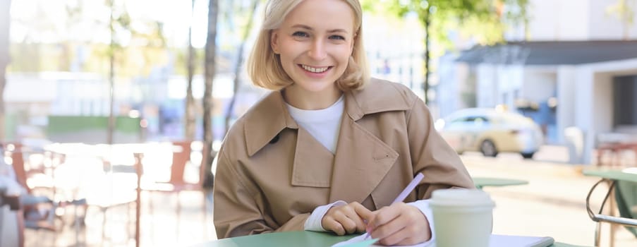 Image of young blonde woman in stylish trench, sitting in outdoor cafe, drinking coffee and working on documents, studying, doing homework in her notebook, smiling at camera.