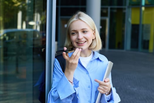 Portrait of beautiful, smiling young student, woman records voice message, talking on speakerphone, standing near office building on street and holding laptop with backpack.