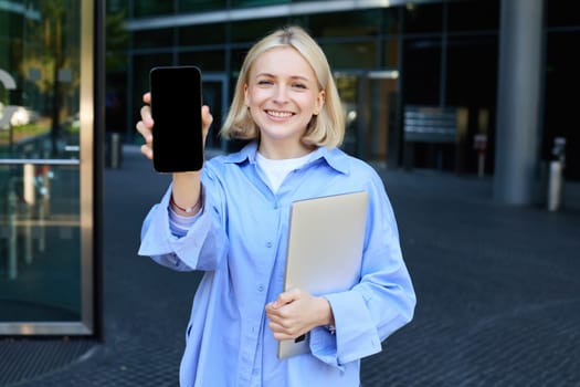 Portrait of beautiful young woman, student posing on street near campus, holding laptop, showing mobile phone screen, demonstrating app or promo on smartphone and smiling.