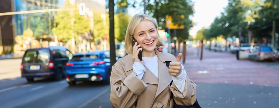 Lifestyle portrait of young blond woman talking on mobile phone, chatting with someone while walking on street, has conversation on smartphone and laughing.