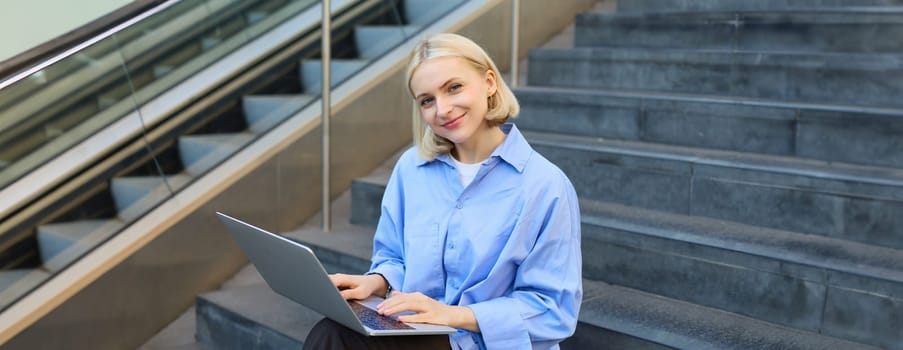 Portrait of woman with laptop, found wifi spot, sitting on stairs outdoors, drinking coffee, working on computer.