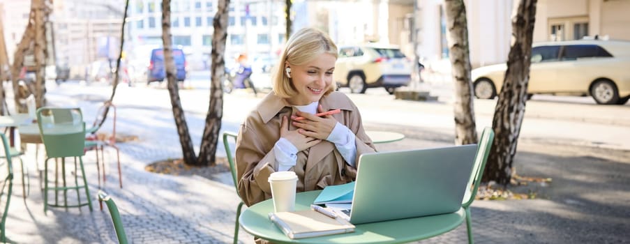 Portrait of young happy woman, looking pleased and surprised at laptop screen, sitting in outdoor cafe, street coffee shop, holding hands on chest and smiling grateful.