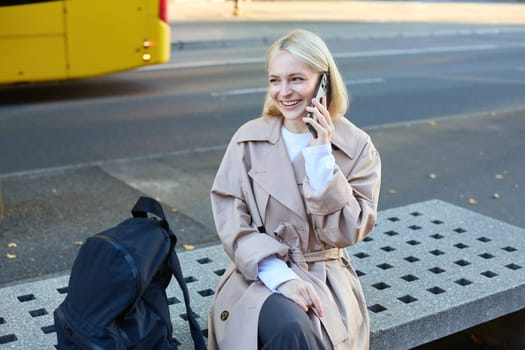 Lifestyle portrait of young beautiful blond girl, sitting on bench outside, talking on mobile phone, chatting with friend and laughing from funny conversation.
