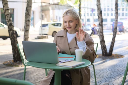 Image of excited blond girl, celebrating, sitting in coffee shop on sunny day, looking at laptop and triumphing. Lifestyle and emotions concept
