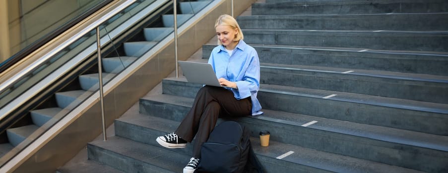 Freelance employee, woman working on her project, using laptop, sitting outdoors on stairs, drinking takeaway coffee.