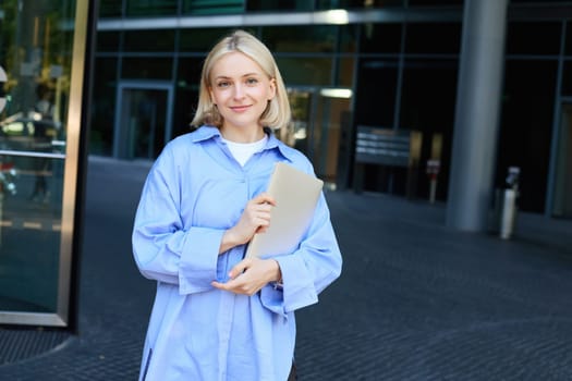 Image of young female employee, working woman standing near her office on street, holding pile of documents and smiling. People and lifestyle concept