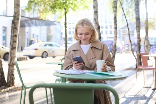 Image of young woman drinking coffee, sitting in outdoor cafe with smartphone, sending message, reading notification on mobile phone and smiling.