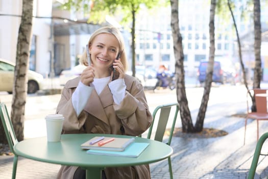 Lifestyle and urban people concept. Young smiling girl, student sitting in cafe outdoors, talking on mobile phone, chatting with friend or coworker over the telephone, studying or doing homework.