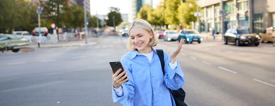 Portrait of young office worker, woman walking along street, chatting on the phone, looking at smartphone and showing smth behind her, video chats, connects to online conversation via mobile app.