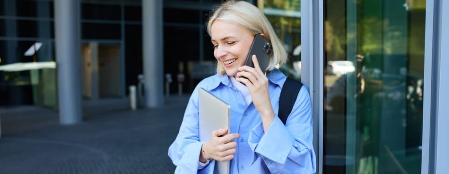 Young modern woman, female employee, standing near office building, answers phone call, talking, holding laptop and backpack, smiling while having a nice, friendly chat.