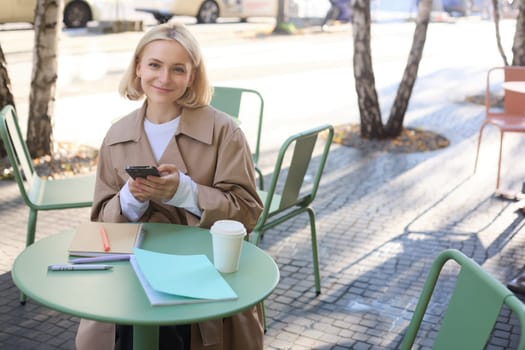 Portrait of smiling young woman using mobile phone while doing homework, working on project, sitting outdoors in cafe, drinking coffee and studying, holding smartphone.