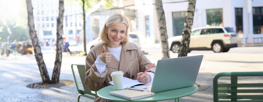 Portrait of beautiful blond female model, showing thumbs up, sitting with laptop and study material in coffee shop, gives approval. Lifestyle concept