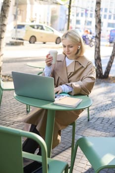 Vertical shot of young blond woman with laptop, sitting with coffee in street cafe, working outdoors on computer, wearing wireless headphones and smiling.