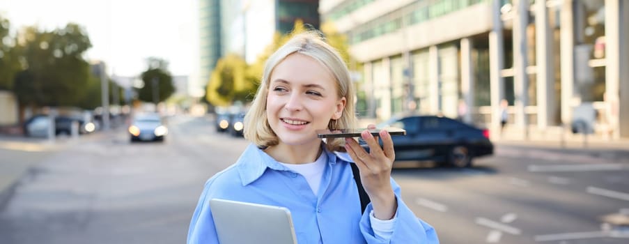 Image of happy young woman walking along street, listening to voice message, holding smartphone speaker near ear to hear better, has laptop in her hand.