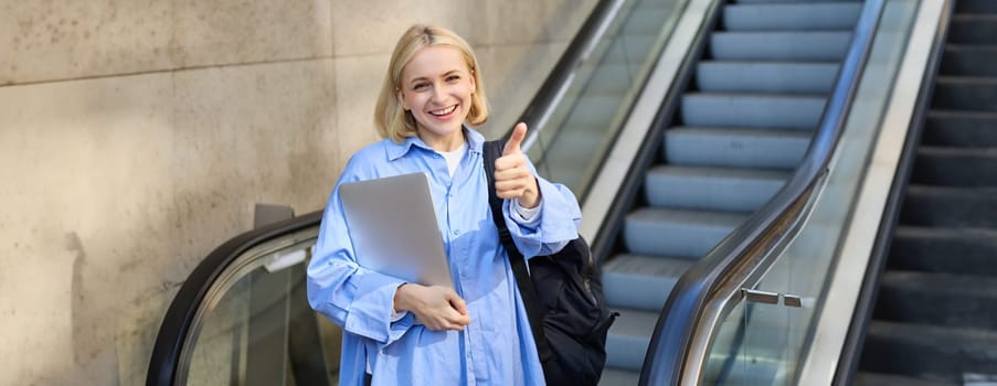 Friendly, smiling young woman, college student with laptop and backpack, using escalator, showing thumbs up in approval, recommending product or company.