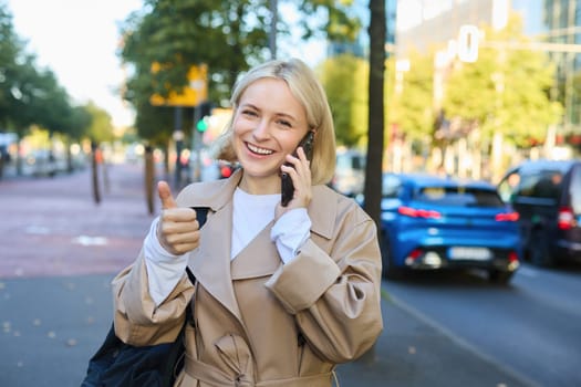 Lifestyle portrait of young blond woman talking on mobile phone, chatting with someone while walking on street, has conversation on smartphone and laughing.