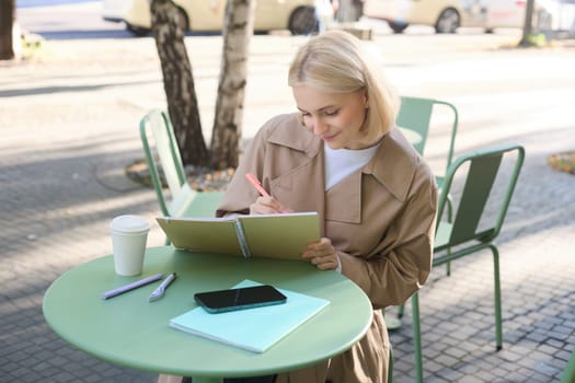 Portrait of beautiful blond woman, sitting in outdoor coffee shop, drawing in cafe in notebook, making sketches outside on street,