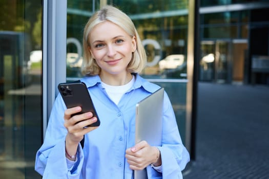 Portrait of young female student standing outside university campus, posing near building, holding laptop and smartphone, wearing blue collar shirt.