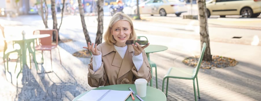 Portrait of young urban female model, sitting in cafe outdoors, recording voice message with upset, confused face, shrugging, holding mobile phone near mouth.