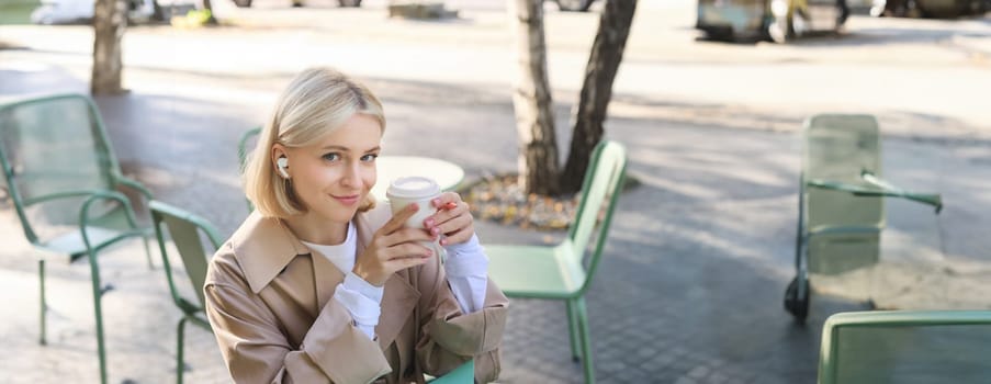 Vertical shot of cute blond woman, sitting in street cafe outdoors, wearing wireless headphones, using laptop, drinking her coffee and smiling at camera.