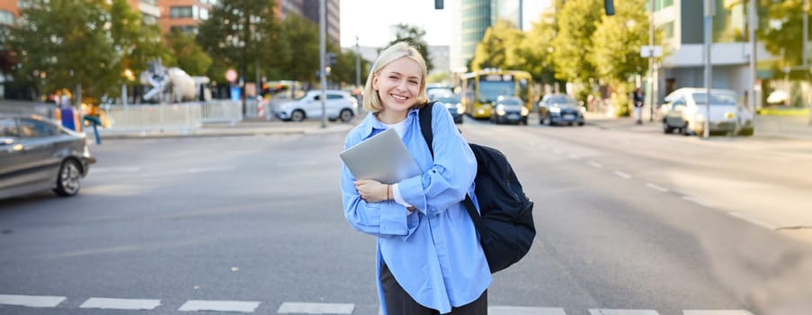 Image of young college student, happy female model on street, posing with backpack and laptop outdoors, with roads and cars behind her back, smiling at camera.