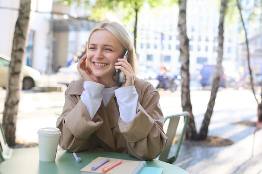 Close up portrait of smiling blond woman, sitting at table outside cafe, talking on mobile phone, having happy, lively chat.