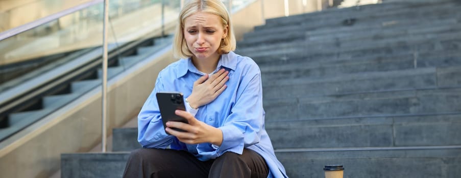 Portrait of young woman looking with pity and compassion at smartphone camera, staring at something heart-breaking on mobile phone, feeling sad and crying, sitting on stairs outdoors.