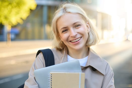 Close up portrait of smiling blond woman, standing on street with notebooks, carries journal and work documents, looking happy at camera.