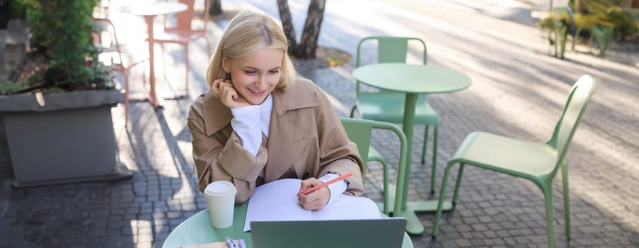 Image of smiling young female student, working on project, sitting outside on sunny day in cafe, looking at laptop and making notes, studying, doing homework outdoors.