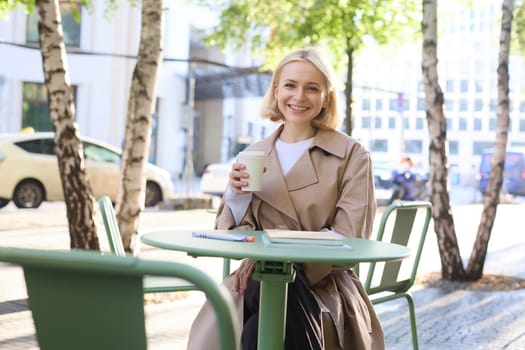 Portrait of young blonde woman sitting in cafe, doing homework and drinking coffee, writiing in journal, using notebook on fresh air.