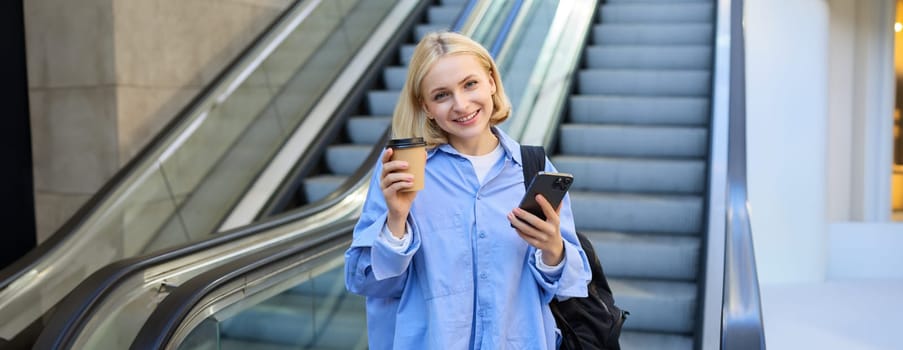 Image of beautiful female model, student standing near escalator, raising cup of takeaway coffee, smiling and looking happy, has mobile phone in hand and backpack on shoulder.