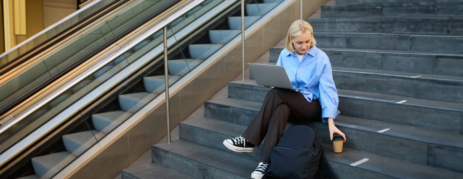Modern young woman, student working on laptop, sitting outside campus in stairs with backpack and takeaway coffee, drinking her beverage and studying.