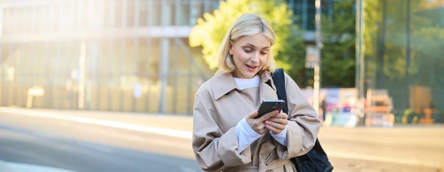 Portrait of surprised girl on street, woman looking at her phone with raised eyebrows, reading a message on smartphone, waiting for someone near the road, holding backpack.