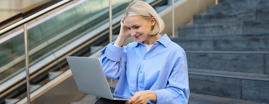 Young woman, student in blue shirt, working on laptop, sitting outdoors on street stairs, working on project online, connects to public wifi, elearning.
