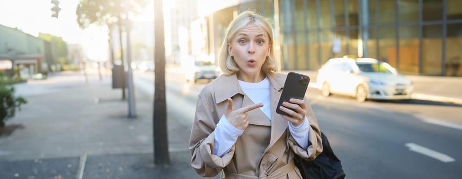 Image of surprised girl on street, pointing at mobile phone and gasping amazed, saying wow. Technology and urban lifestyle concept