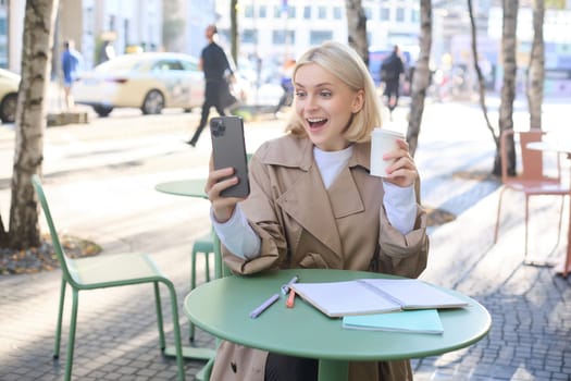 Image of blond female model, looks surprised at her social media account, staring at smartphone with amazed face, drinking coffee outdoors, sitting outside.