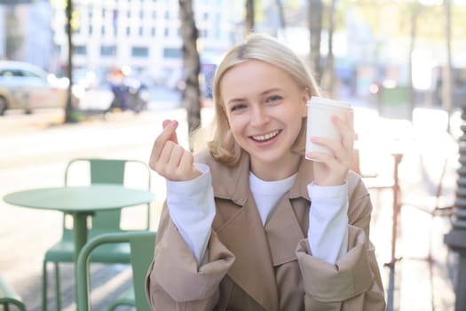 People and leisure concept. Happy blonde girl, sitting with cup of coffee in street cafe, showing finger heart sign, enjoys spending time outdoors.