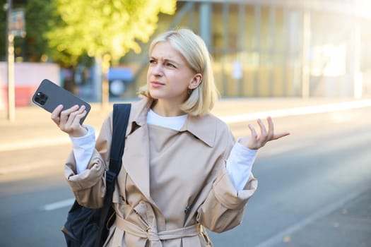 Portrait of woman with confused face, walking on street, holding smartphone, feeling lost, using mobile map application, shrugging shoulders.