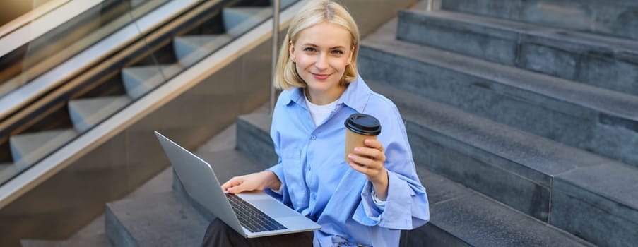 Image of stylish young woman, college student with laptop, sitting on city stairs and drinking coffee, working on project, studying outdoors, connects to public wifi, enjoying weather.