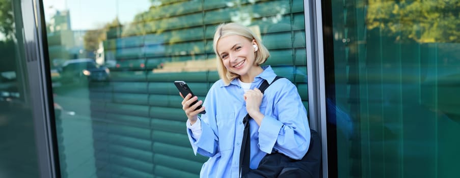 Portrait of beautiful blond girl, smiling woman in wireless earphones, listening to music on smartphone, waiting for friend on street, standing outdoors, leaning on building.