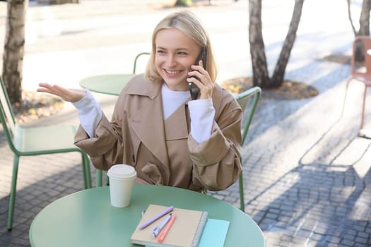 Close up portrait of happy, charismatic young woman talking on mobile phone, sitting in cafe alone, outside on sunny day, answer a call, having friendly conversation.