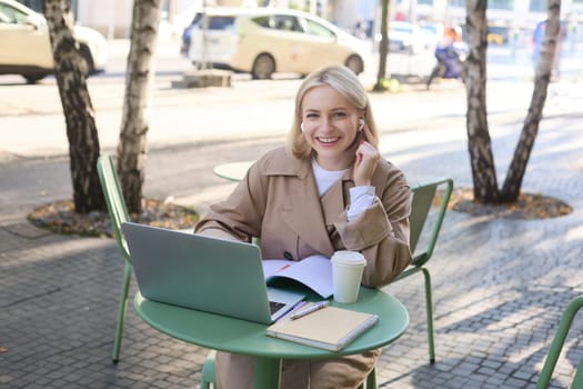 Portrait of beautiful blond woman, wearing wireless headphones, using laptop, studying in outdoor coffee shop, making notes, working on project.