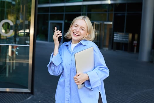 Carefree blond woman with laptop and smartphone, singing, listening music on mobile phone app, posing near office building or college campus on street.