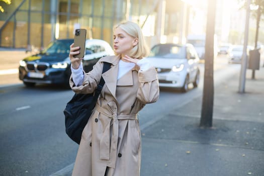 Portrait of beautiful young woman, social media blogger, taking selfie on smartphone while posing on city road, pucker lips, kissing face, using mobile phone camera.