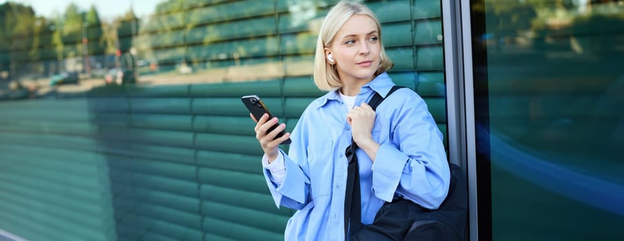 Lifestyle portrait of smiling young female model, student with backpack, waiting for someone on street, standing outdoors with smartphone.